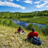 Zwei Wanderer liegen gemütlich im Gras und blicken auf einen Wald und einen Fluss